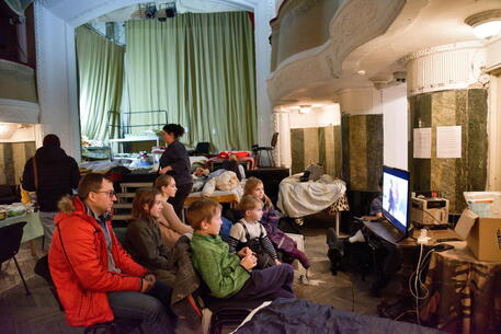 Una famiglia guarda la tv in un teatro trasformato in campo profughi in Ucraina © EPA