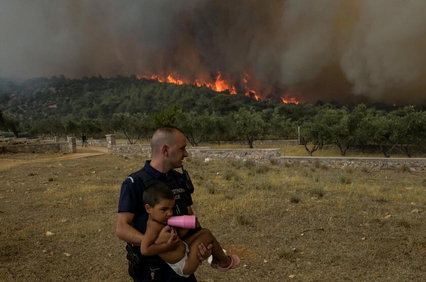 Un poliziotto evacua un bambino da un incendio nel villaggio di Agios Charalampos, vicino ad Atene © ANSA/AFP
